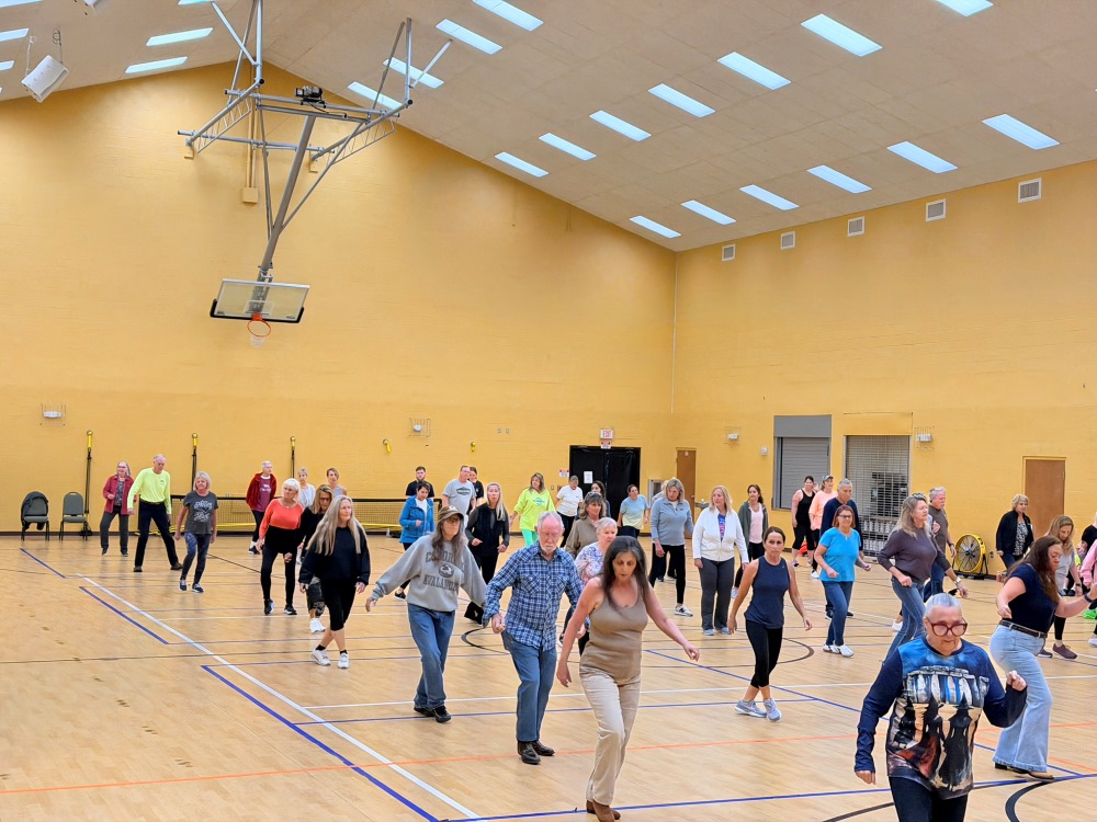 Large group of adults follows a line dance routine in a brightly lit gymnasium